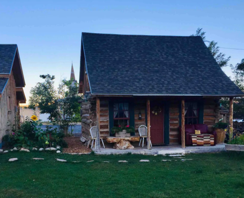 A cozy log cabin with a sloped roof, surrounded by greenery and a small garden, set against a clear blue sky.